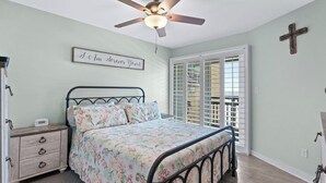Um quarto com cama, ventoinha no teto, cômoda, mesa de cabeceira, relógio na parede, janela com persianas e uma cruz na parede.