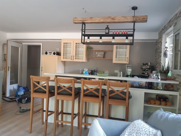 A modern kitchen with wooden bar stools, a built-in coffee machine, and a wooden ceiling light fixture.
