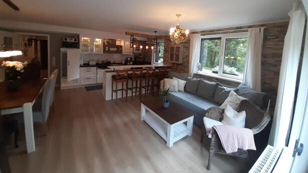 Ein großzügiger Wohnbereich mit Sofa, Couchtisch und Esstisch. Im Hintergrund ist eine Küche mit Steinwand und einer Chandelier zu sehen.