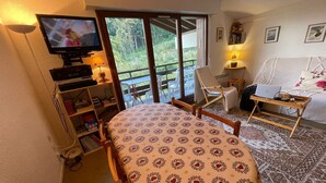 Ein gemütliches Zimmer mit einem Bett, einem Fernseher, einem Laptop auf einem Nachttisch, einem Sessel und Blick ins Grüne.
