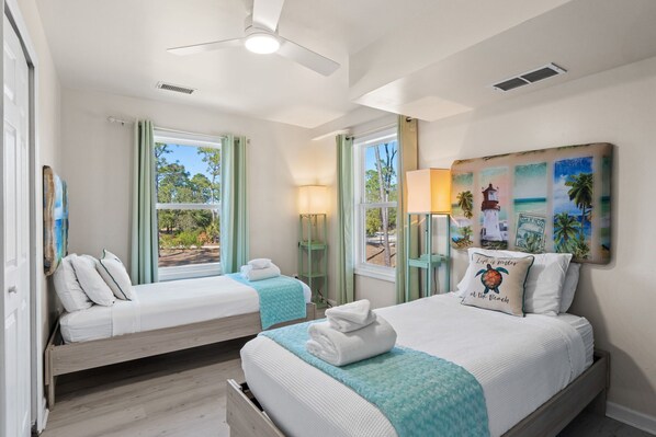 A hotel room with two beds, a ceiling fan, a large window with curtains, a beach-themed wall art, and a lamp.
