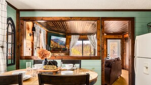 A rustic cabin interior with a wooden dining table, a sofa, and a large window with a painting.
