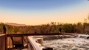 A hot tub area with a wooden deck, a chair, and a view of a forested landscape.