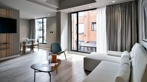 Ein modernes Wohnzimmer mit einer Couch, einem Essbereich mit Stühlen, einem an der Wand montierten Fernseher und großen Fenstern mit Vorhängen.
