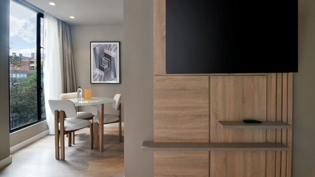 A modern living room with a large window, a flat-screen TV mounted on a wooden panel wall, a dining area with a glass table and chairs, and a framed abstract artwork on the wall. - w Cozy 1BR w/ Brilliant Balcony in Unicentro (Bogotá)