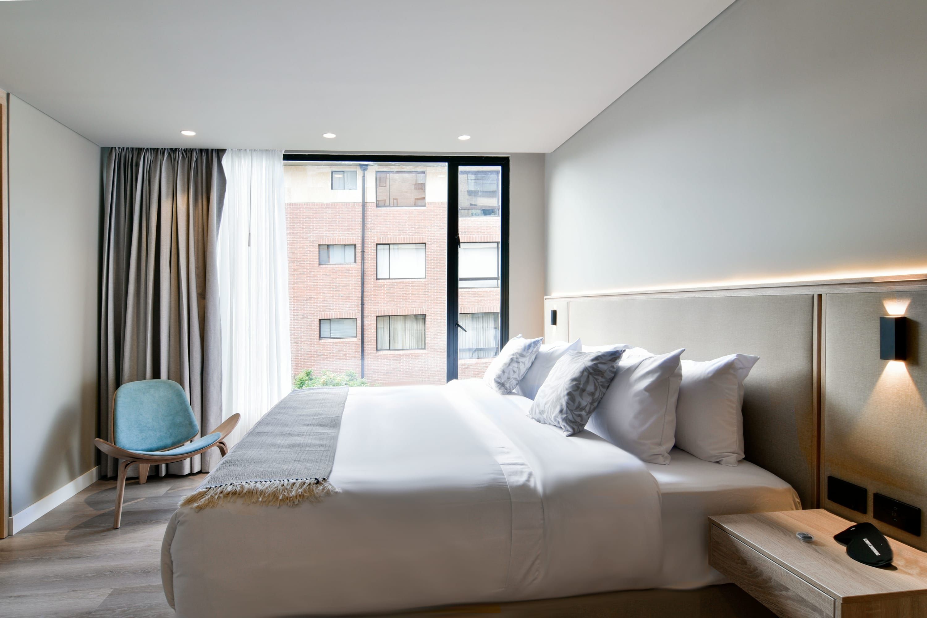 Une chambre d’hôtel moderne avec un grand lit, une chaise, une table de chevet et une vue sur un immeuble en briques par la fenêtre.