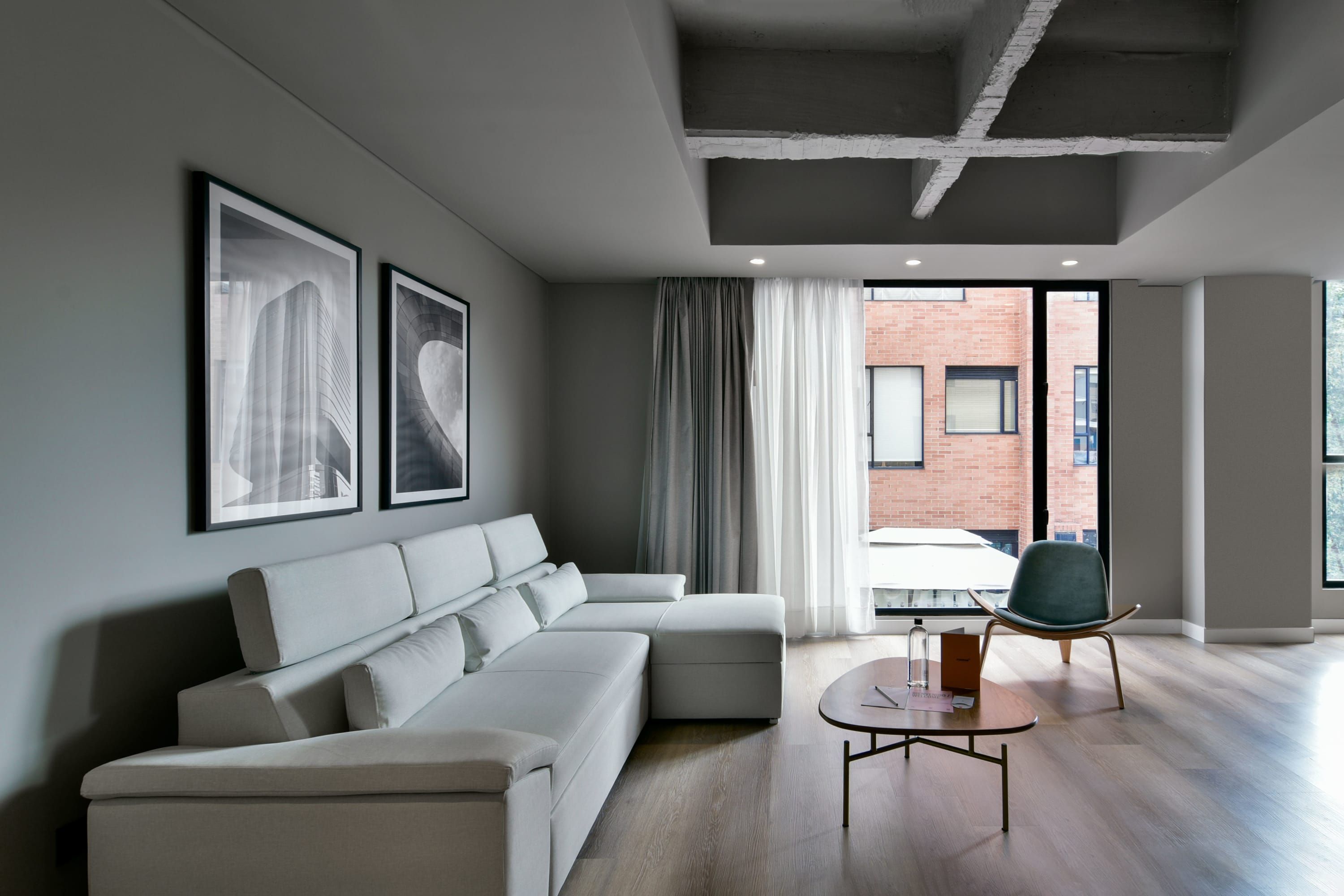 Una sala de estar moderna con un sofá modular, una mesa de centro redonda y una silla. Hay dos cuadros enmarcados en la pared, una ventana con cortinas y se ve una pared de ladrillo a través de la ventana.