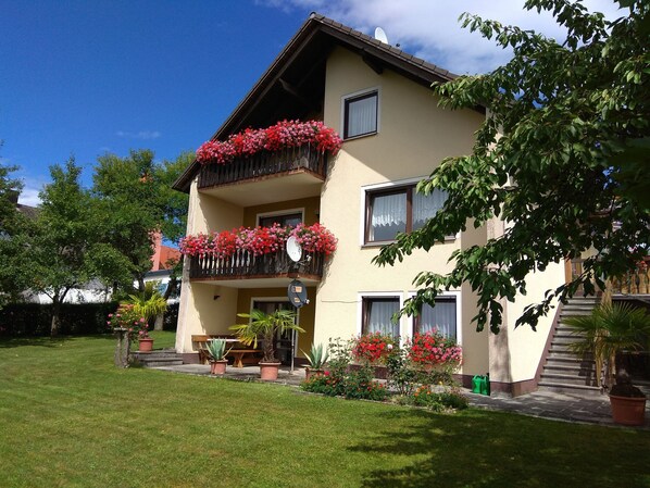 A two-story building with balconies adorned with red flowers, a small garden area with potted plants, and a clear blue sky.