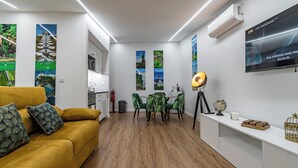 Un salon moderne avec un canapé jaune, un coin repas avec des chaises vertes, un téléviseur fixé au mur et un meuble blanc.