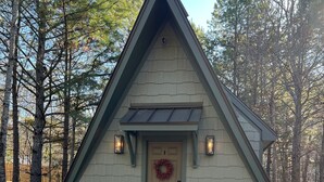 A small, triangular-shaped cabin with a wooden door and a wreath, surrounded by trees.