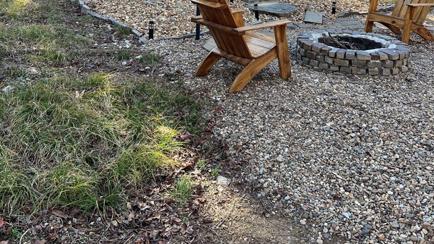 A cabin with a sloped roof, a front door with a wreath, and a fire pit area with two wooden chairs.
