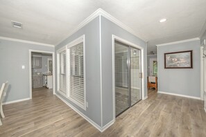 Un interno moderno con una cucina sullo sfondo, un corridoio con una porta in vetro e un quadro appeso al muro.