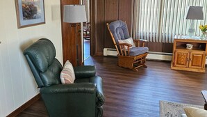 A living room with a dark green leather armchair, a wooden rocking chair, and a small wooden cabinet with a vase of flowers.