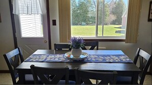 A dining room with a table set for six, a potted plant on the table, and a view of a grassy yard through a large window.