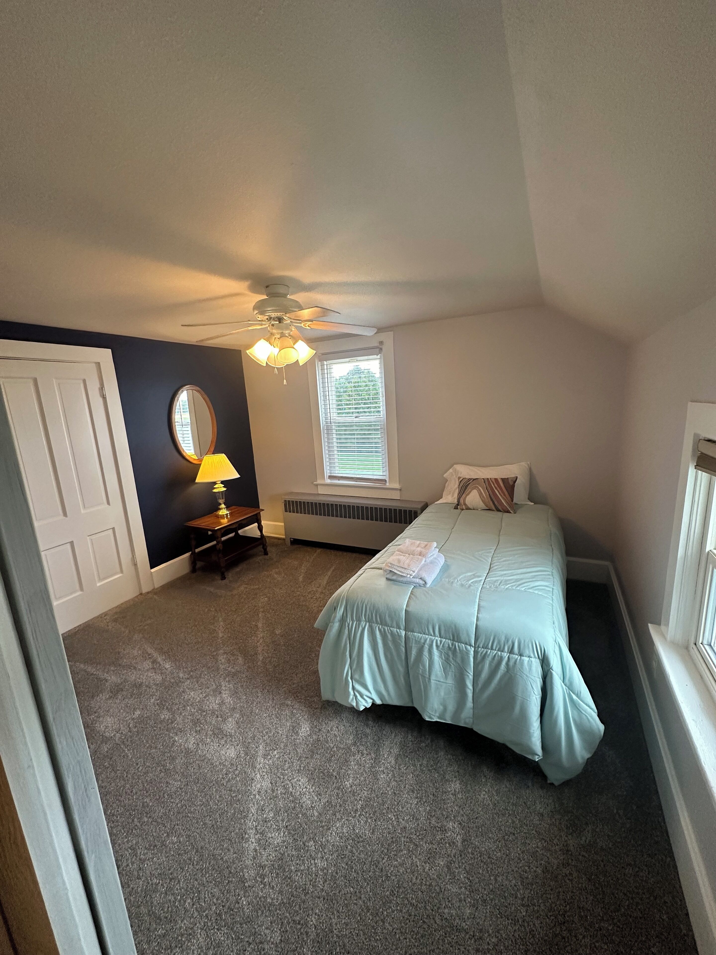 A bedroom with a bed, a ceiling fan, a window with blinds, a radiator, and a mirror on the wall.