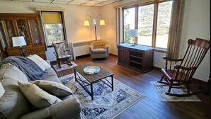 A living room with a sofa, a coffee table, a chair, a desk, and a rocking chair. There is a window with curtains, a lamp, and a cabinet.