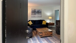 Una sala de estar con una puerta oscura, un sofá azul con cojines amarillos, una mesa de centro de madera y un cuadro en la pared.