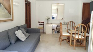 A compact living space with a grey sofa, a dining table with four chairs, and a kitchen area in the background. - D&P Suites 2 Salemi (Salemi)