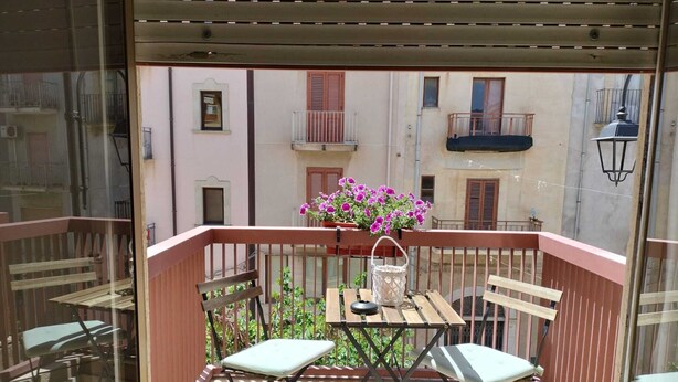A balcony with a table and two chairs, a potted plant with flowers, and a view of a building with balconies. - D&P Suites 2 Salemi (Salemi)