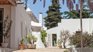 Un edificio blanco con tejado de tejas, rodeado de macetas con plantas y una pérgola con flores colgantes.