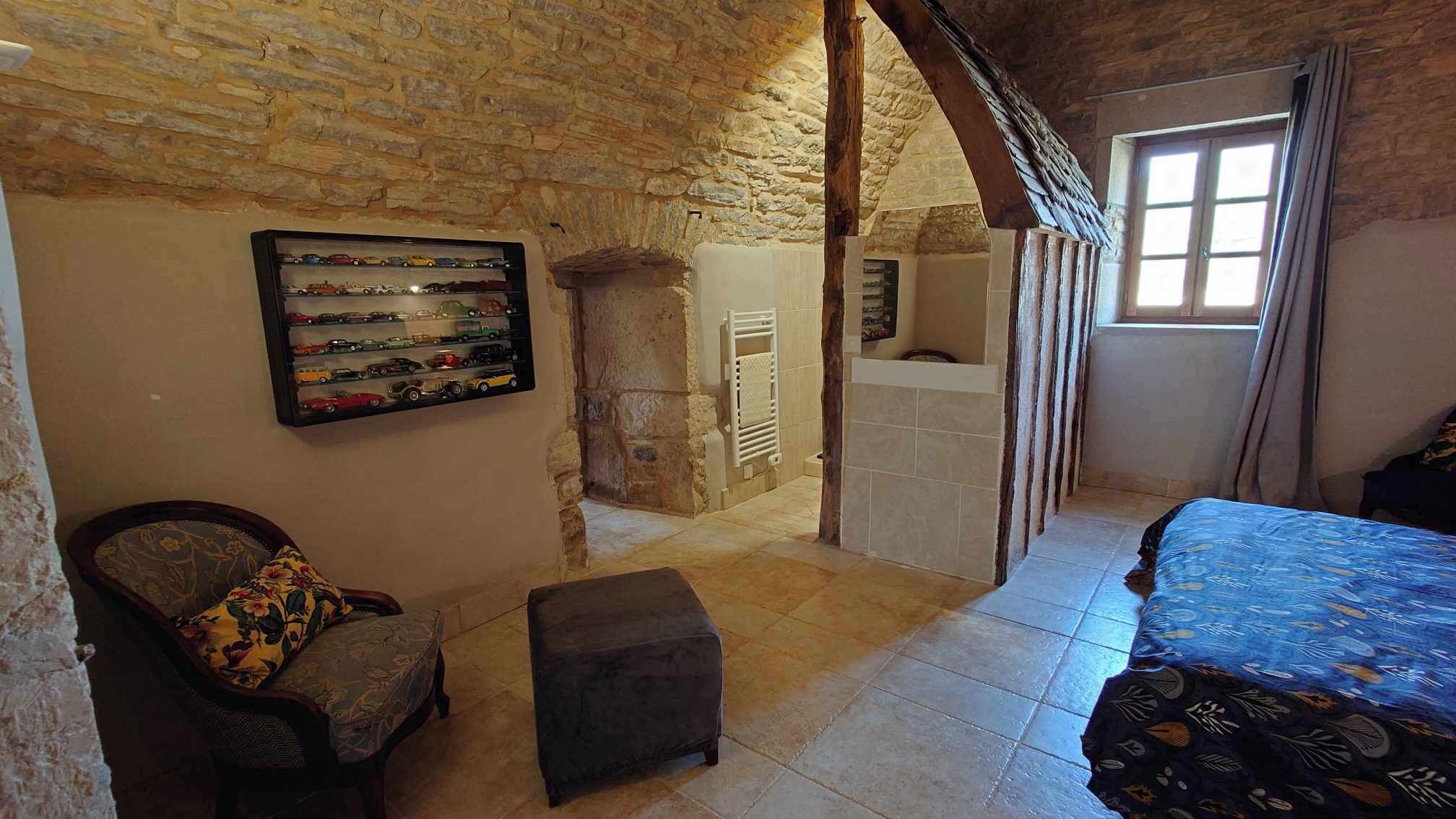 Un dormitorio con pared de piedra, cama con colcha estampada, sillón con cojín floral, un pequeño reposapiés y una exhibición de coches de juguete en la pared.