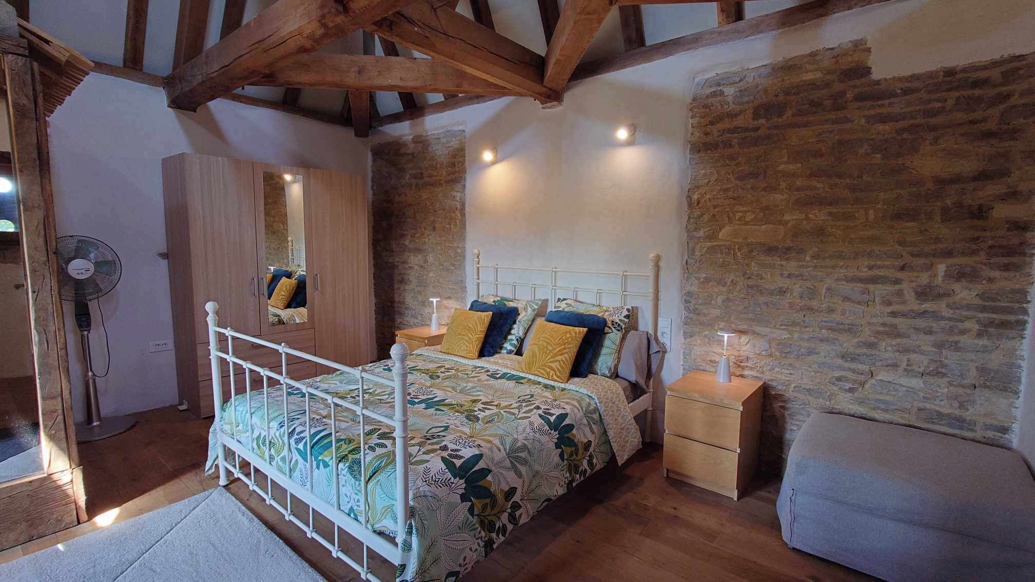 A bedroom with a wooden ceiling, a bed with floral bedding, a nightstand, a mirror, and a fan.