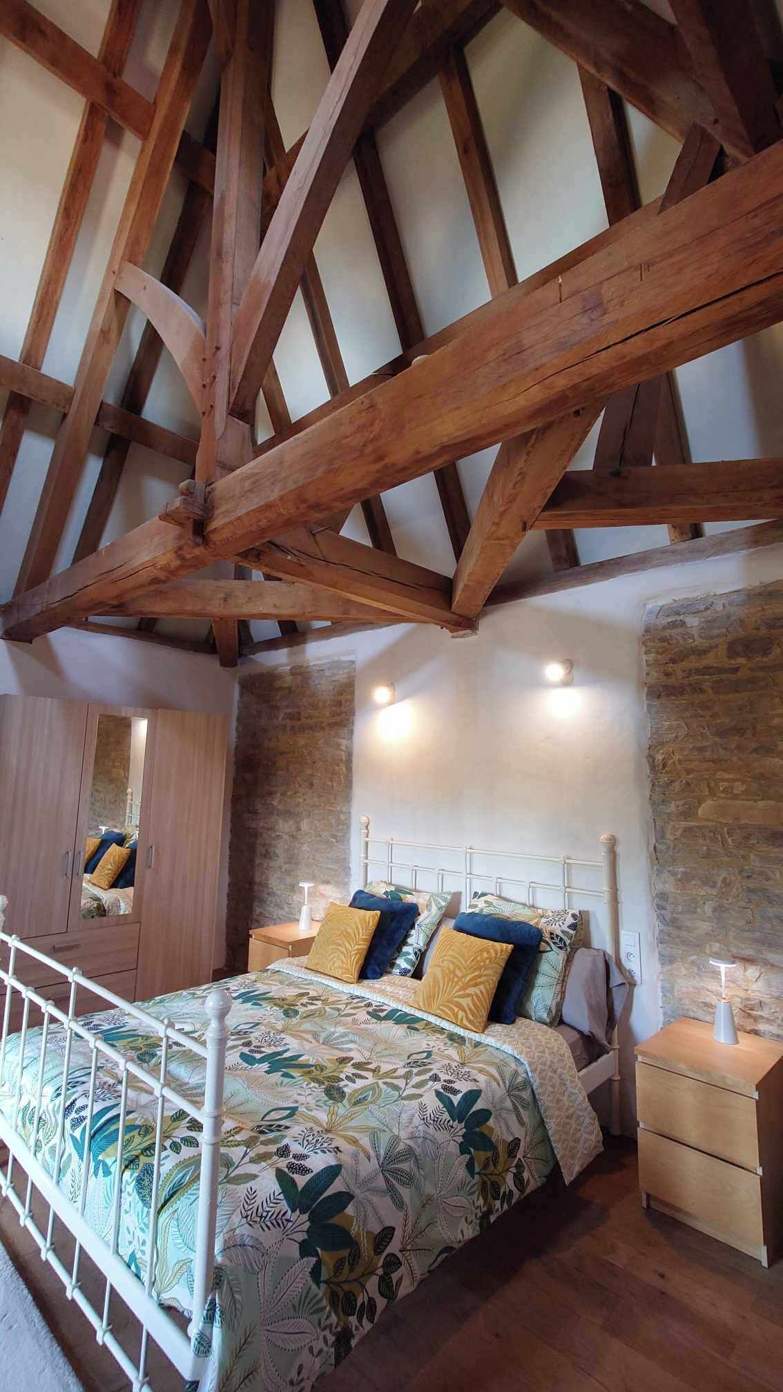 Een slaapkamer met een hoog plafond, zichtbare houten balken, een bed met bloemenbeddels, een nachtkastje en een kledingkast.