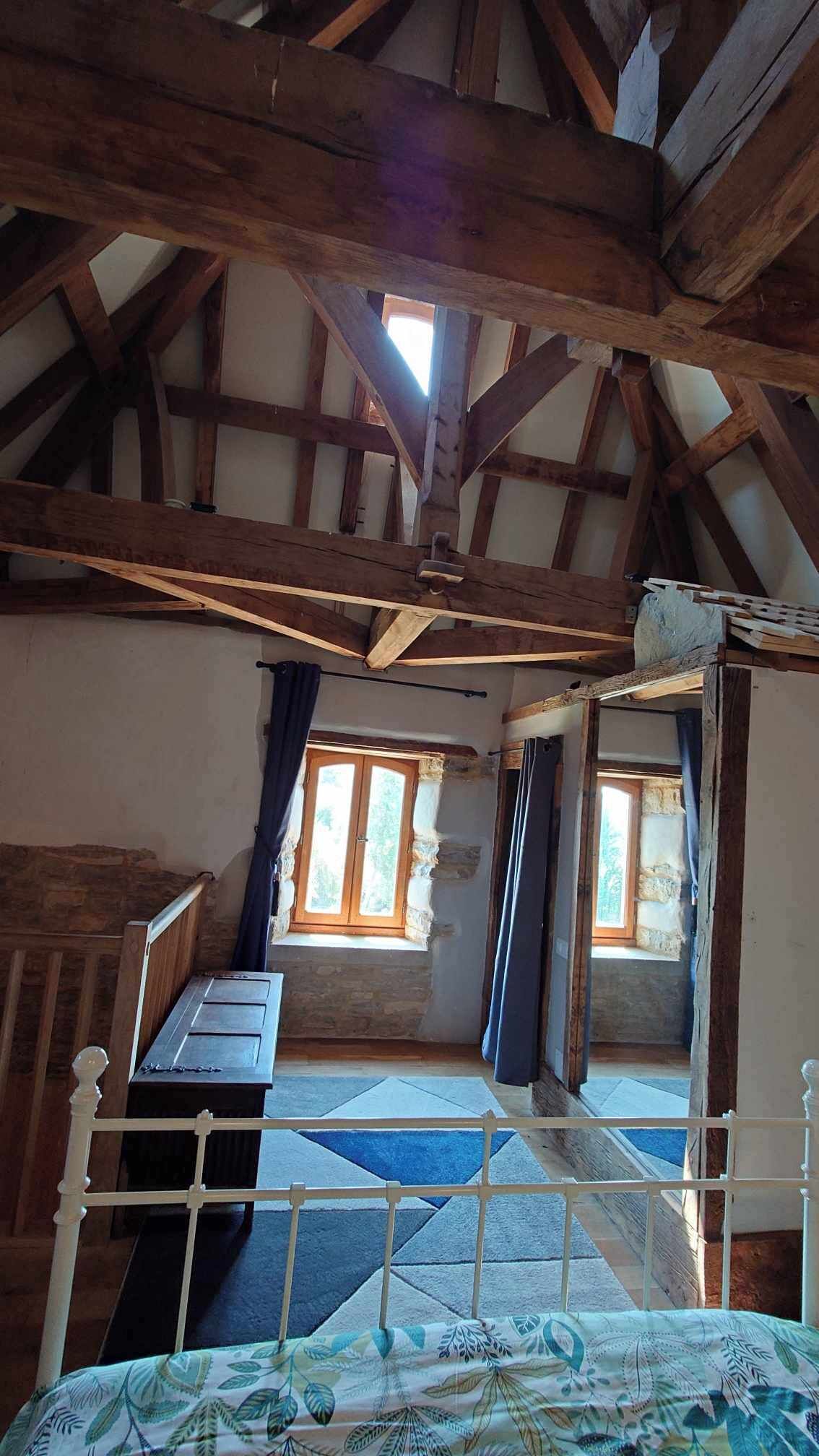 A rustic room with exposed wooden beams, a bed with a patterned bedspread, a window with curtains, and a stone wall.