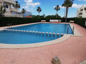 Une piscine dont le bord est revêtu de carreaux, entourée d’un chemin pavé en briques, de palmiers et d’un bâtiment doté de balcons.