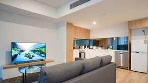 A modern living room with a grey sofa, a flat-screen TV on a wooden stand, and a small glass table. A kitchen with white cabinets and stainless steel appliances is visible in the background. - Charming CBD Escape - 1BR Apartment with Pool & Gy (Adelaide)