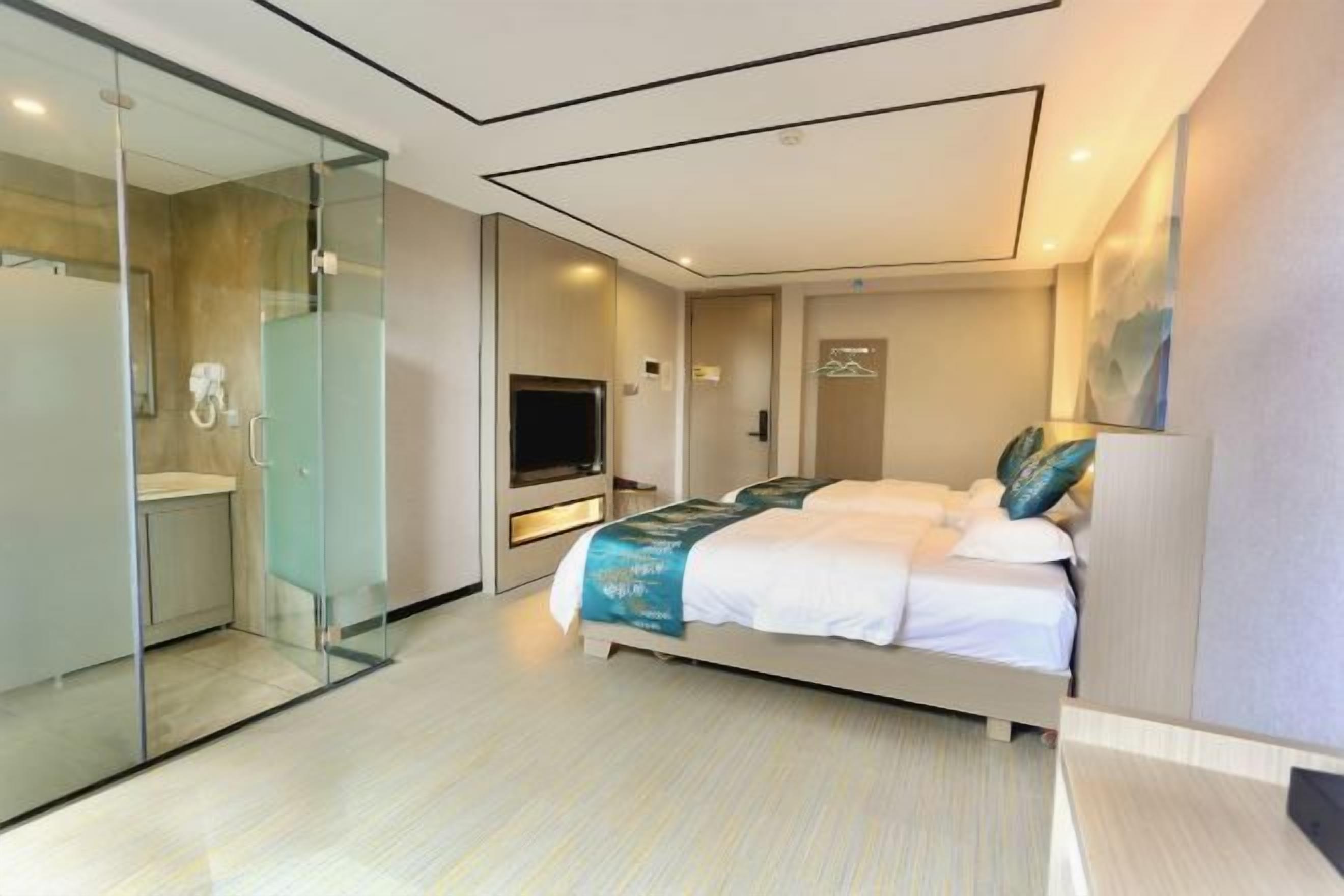 A modern hotel room with a glass-enclosed shower, a bed with a blue and white bedspread, a flat-screen TV, and a small desk area.