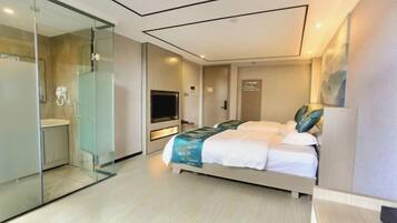 A modern hotel room with a glass-enclosed shower, a bed with a blue and white bedspread, a flat-screen TV, and a small desk area.