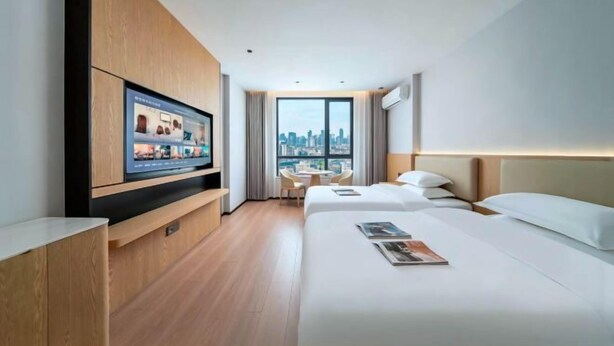 A modern hotel room with a large TV, two beds, a dining area, and a city view.