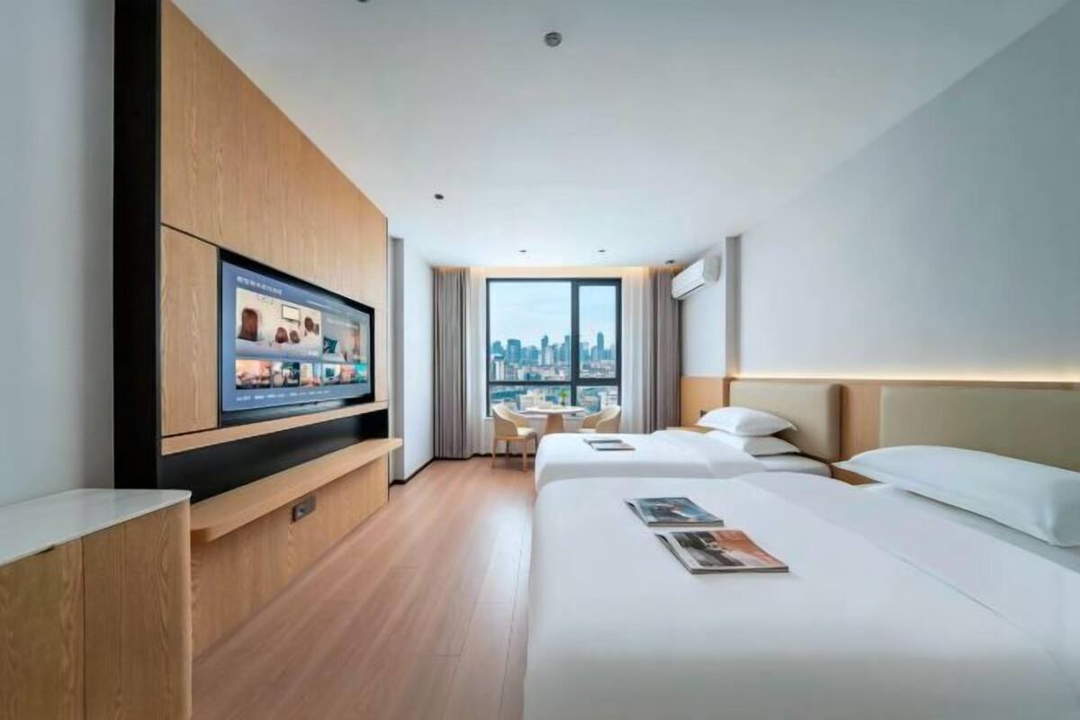 Ein modernes Hotelzimmer mit großem Fernseher, zwei Betten, einer Essecke und Blick auf die Stadt.