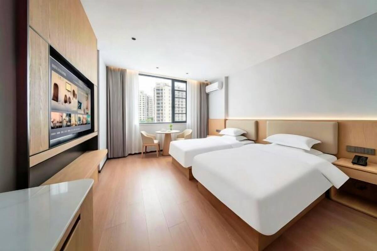 Ein Hotelzimmer mit Fernseher, zwei Betten, einem Schreibtisch, einem Stuhl und einem Fenster mit Blick auf die Stadt.
