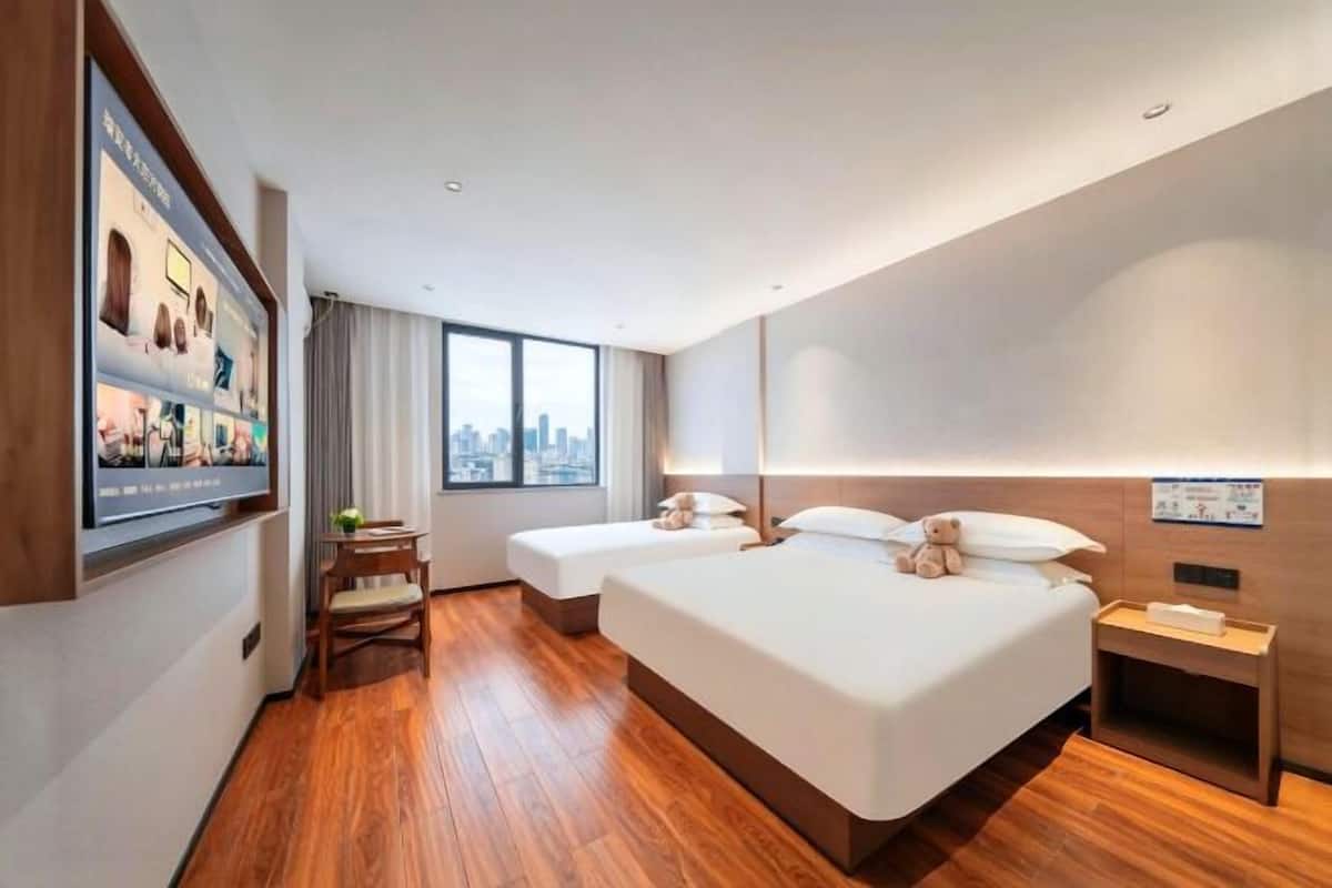 Ein Hotelzimmer mit einem großen Bett, einem kleineren Bett, einem Schreibtisch, einem Sessel, einem Fenster mit Blick auf die Stadt und einem an der Wand befestigten Fernseher.