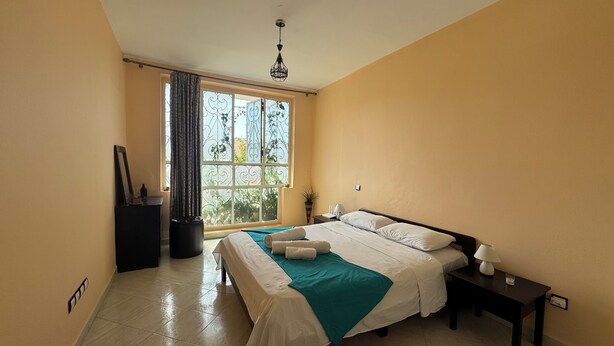 A hotel room with a bed, a nightstand, a lamp, and a window with curtains. - Appartement à 10min des Plages et Souks. Amis à 4 Pates Acceptés ! (Agadir)