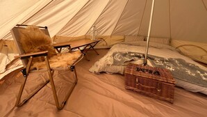 A tent interior with a foldable chair, a small table, a lantern, a basket, and a bed with patterned blankets. - Fossewold Camping Bell Tent 1 of 4 (Blockley)
