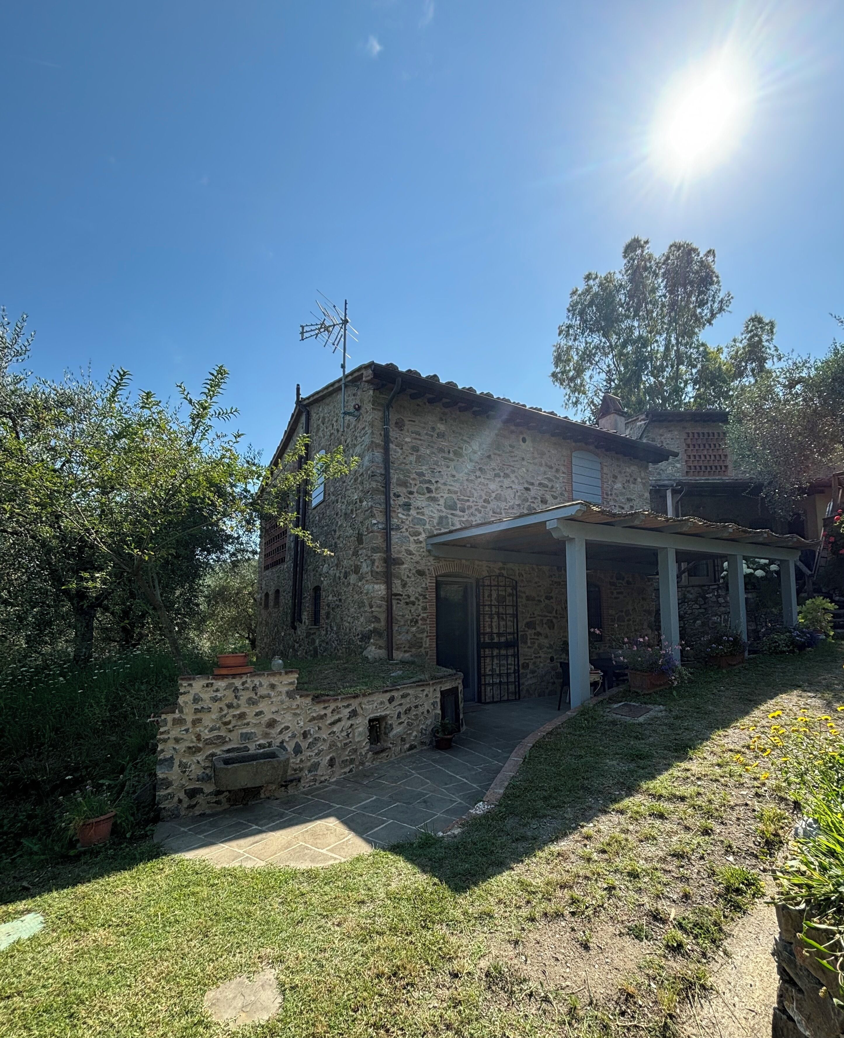 Una casa de piedra con un porche cubierto, rodeada de vegetación y un cielo despejado.