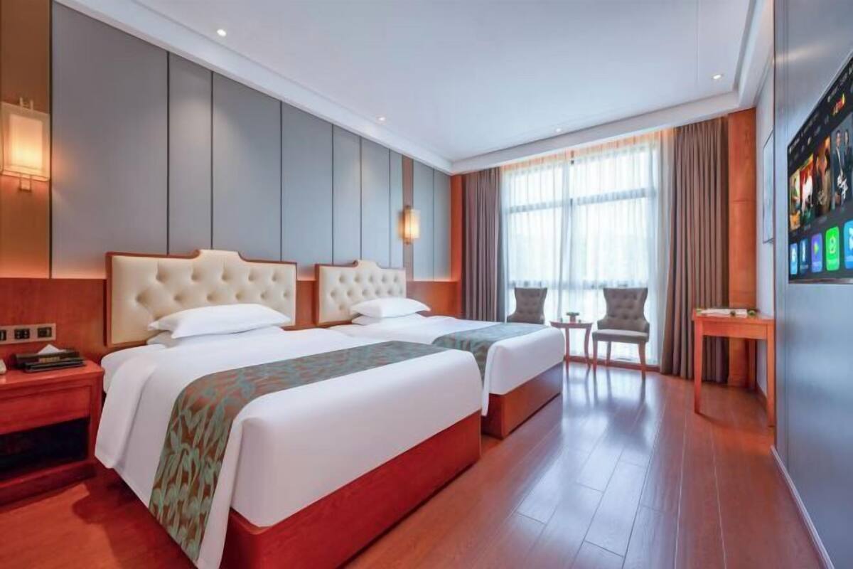 Ein Hotelzimmer mit zwei Betten, einem kleinen Tisch, einem Sessel und einem an der Wand befestigten Fernseher.
