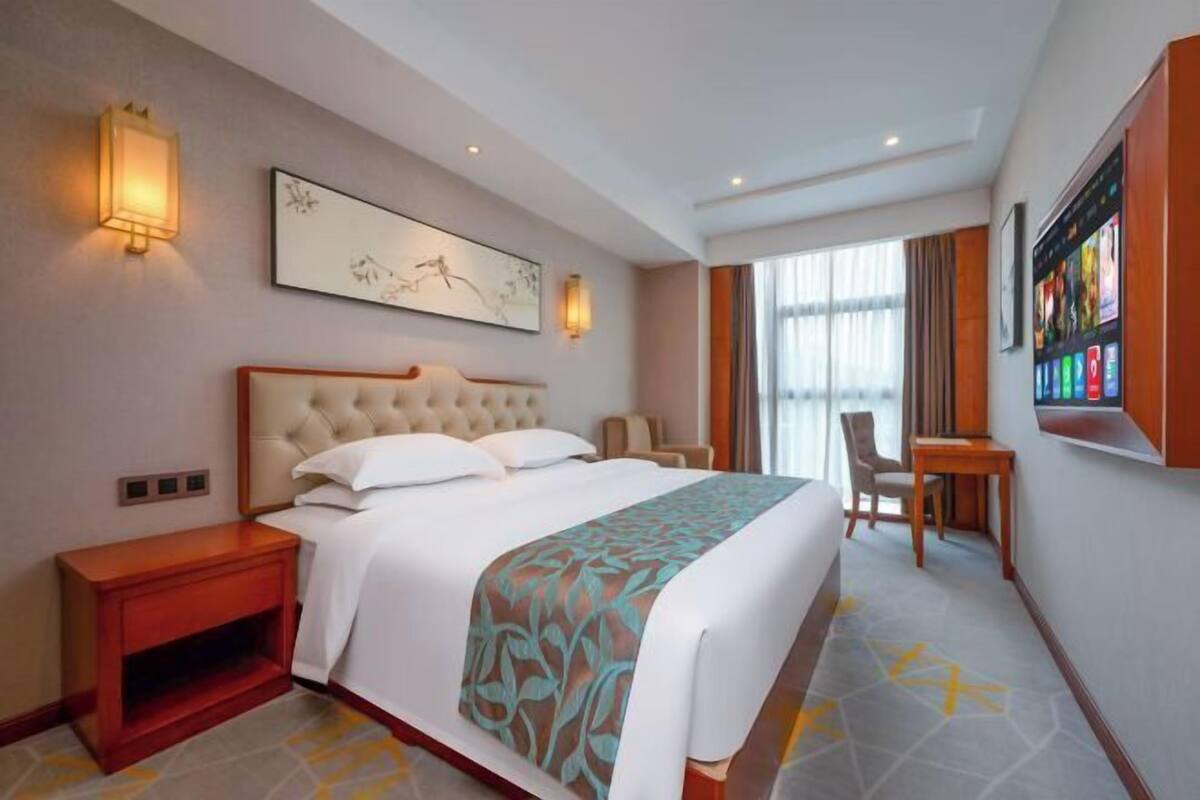 Ein Hotelzimmer mit einem großen Bett, Nachttischen, einem Schreibtisch und einem großen Fenster.