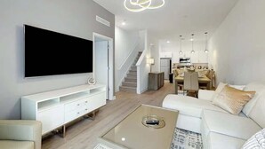 Una sala de estar moderna con un televisor de pantalla plana, muebles blancos, una mesa de centro de vidrio y una escalera que lleva a un comedor.