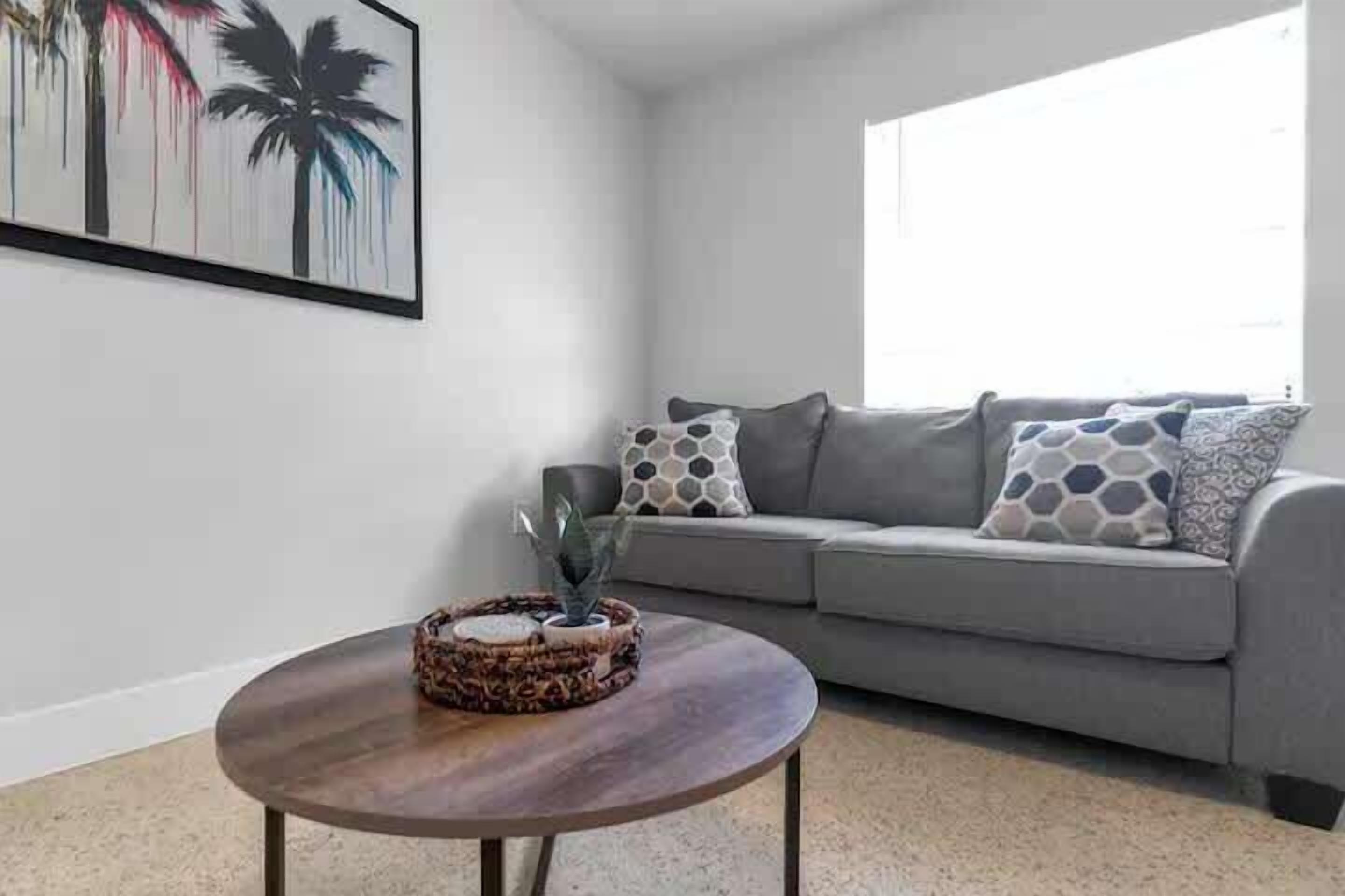 Ein Wohnzimmer mit grauem Sofa, gemusterten Kissen, einem runden Couchtisch mit einer Dekoration und einem Wandbild von Palmen.