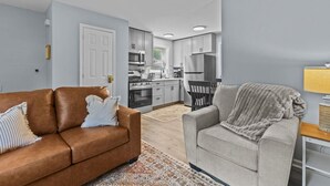 Un salon avec un canapé en cuir marron et un fauteuil gris, une cuisine équipée d’appareils électroménagers modernes et un tapis à motifs sur le sol.