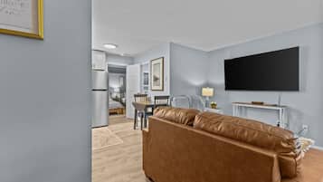 A modern living room with a brown leather sofa, a flat-screen TV mounted on the wall, and a dining area in the background.