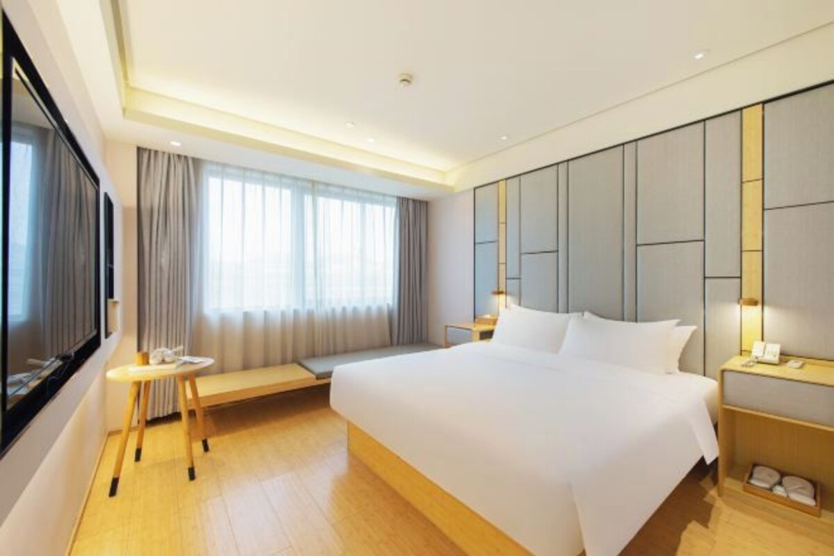 A modern hotel room with a large bed, bedside tables, a small seating area, and a large window with curtains.