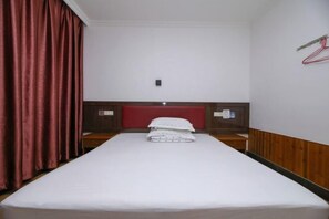 Un lit simple avec un matelas et des oreillers blancs, une tête de lit en bois, une table de chevet en bois et un rideau rouge.