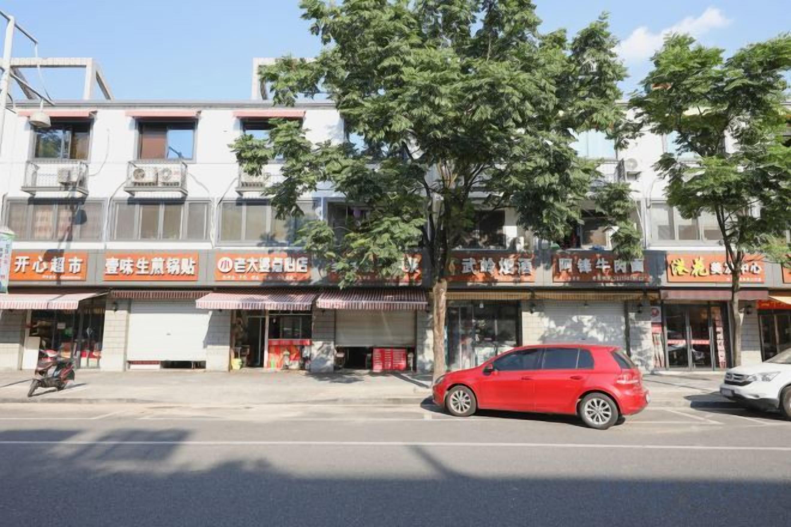 Vista de la calle con una hilera de comercios, un coche rojo estacionado en la acera y un árbol en primer plano.