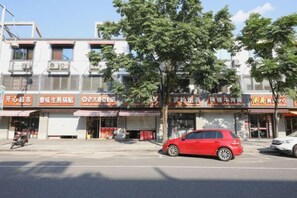 A street view with a row of storefronts, a red car parked on the side, and a tree in the foreground. - Ala Hotel (Ningbo)