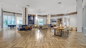 Un hall d’hôtel spacieux, doté d’un parquet en chevrons, d’un espace salon central et de grandes fenêtres.